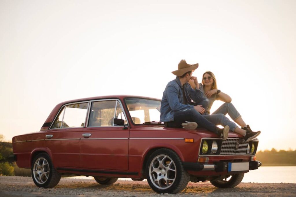 A man and a woman sitting on the hood of a vintage red car at sunset.