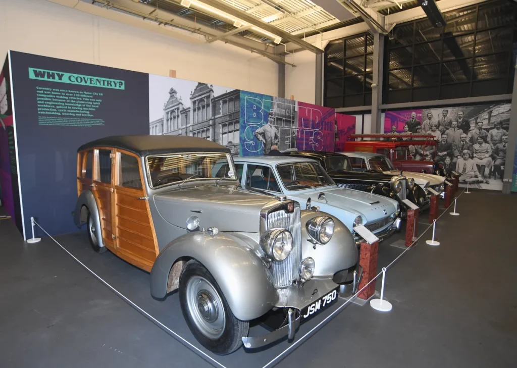 Exhibition hall displaying vintage cars, including a prominent wood-paneled station wagon and a classic silver saloon