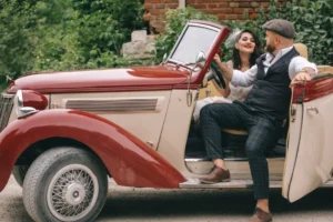 A couple in vintage wedding attire sits inside a classic red and cream convertible car