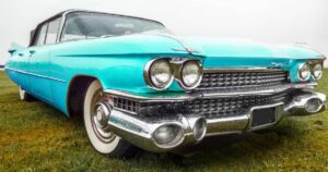 Vintage turquoise car with chrome detailing parked on a grassy field, showing a misty backdrop.