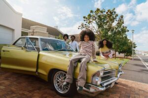 Two adults and two children posing with a vintage yellow car in a suburb.