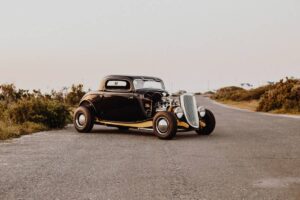 A vintage black hot rod with exposed engine parked on a roadside, surrounded by natural landscape during dusk.