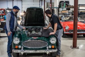 Two men examining the engine of a classic green car in a workshop with other vintage cars visible.