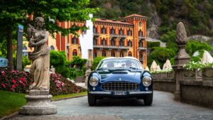 A vintage blue sports car parked on a cobblestone driveway near a classical statue and colorful town buildings.